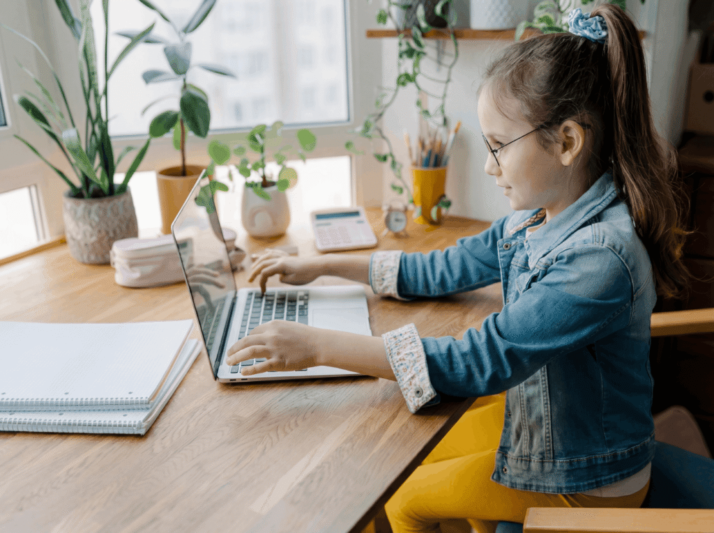Menina usando óculos e jaqueta jeans digitando em um notebook sobre mesa de madeira, com cadernos e plantas ao fundo, em ambiente iluminado por luz natural.