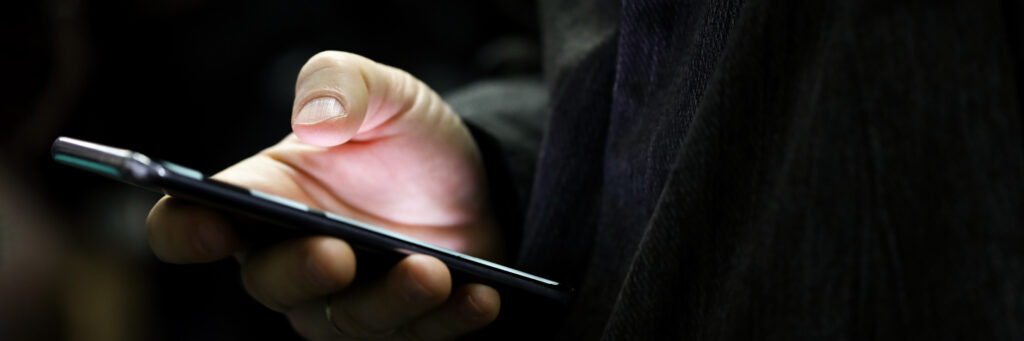 Close-up de uma mão segurando um smartphone em ambiente escuro, com o dedo prestes a tocar na tela, representando riscos e atenção necessária na segurança de links de marketing ao acessar conteúdos digitais.