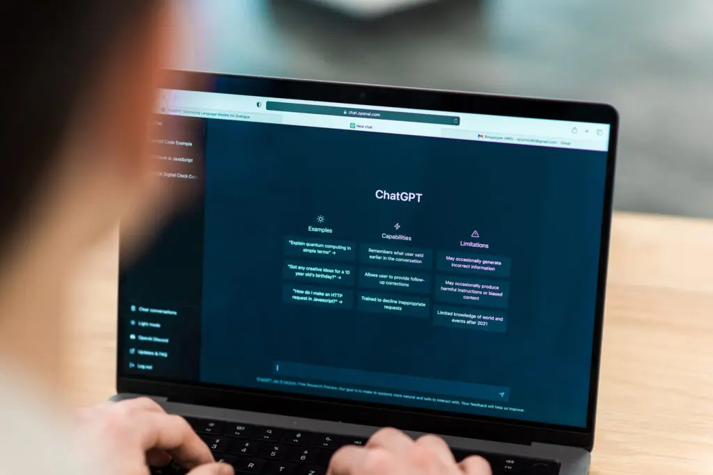 A imagem mostra um notebook aberto com a interface do ChatGPT em destaque na tela, enquanto uma pessoa digita no teclado em primeiro plano, levemente desfocada, direcionando o foco para a interação com a tecnologia; na tela, aparecem seções como “Examples”, “Capabilities” e “Limitations”, reforçando o uso cotidiano de uma IA generativa em um contexto de trabalho ou estudo, e é nesse cenário que Segurança de Dados em IAs Generativas se torna central, ao evidenciar que, por trás da simplicidade e eficiência da interface, existe uma camada crítica relacionada aos dados inseridos, seu processamento e os potenciais riscos envolvidos nessa interação.